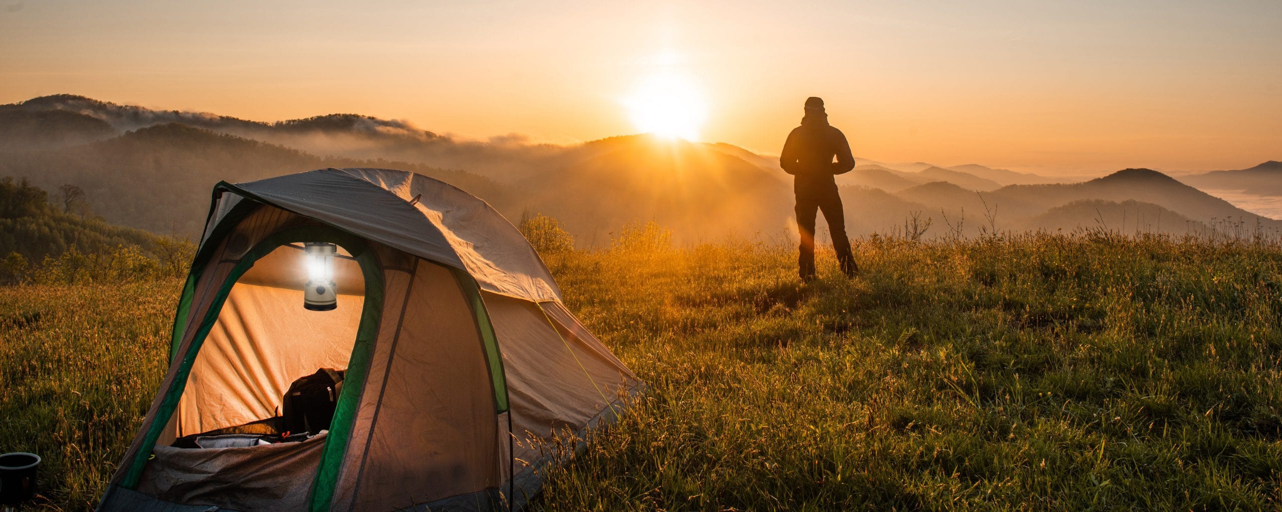 Mann sieht sich einen Sonnenaufgang an, in seinem Zelt leuchtet eine Laterne